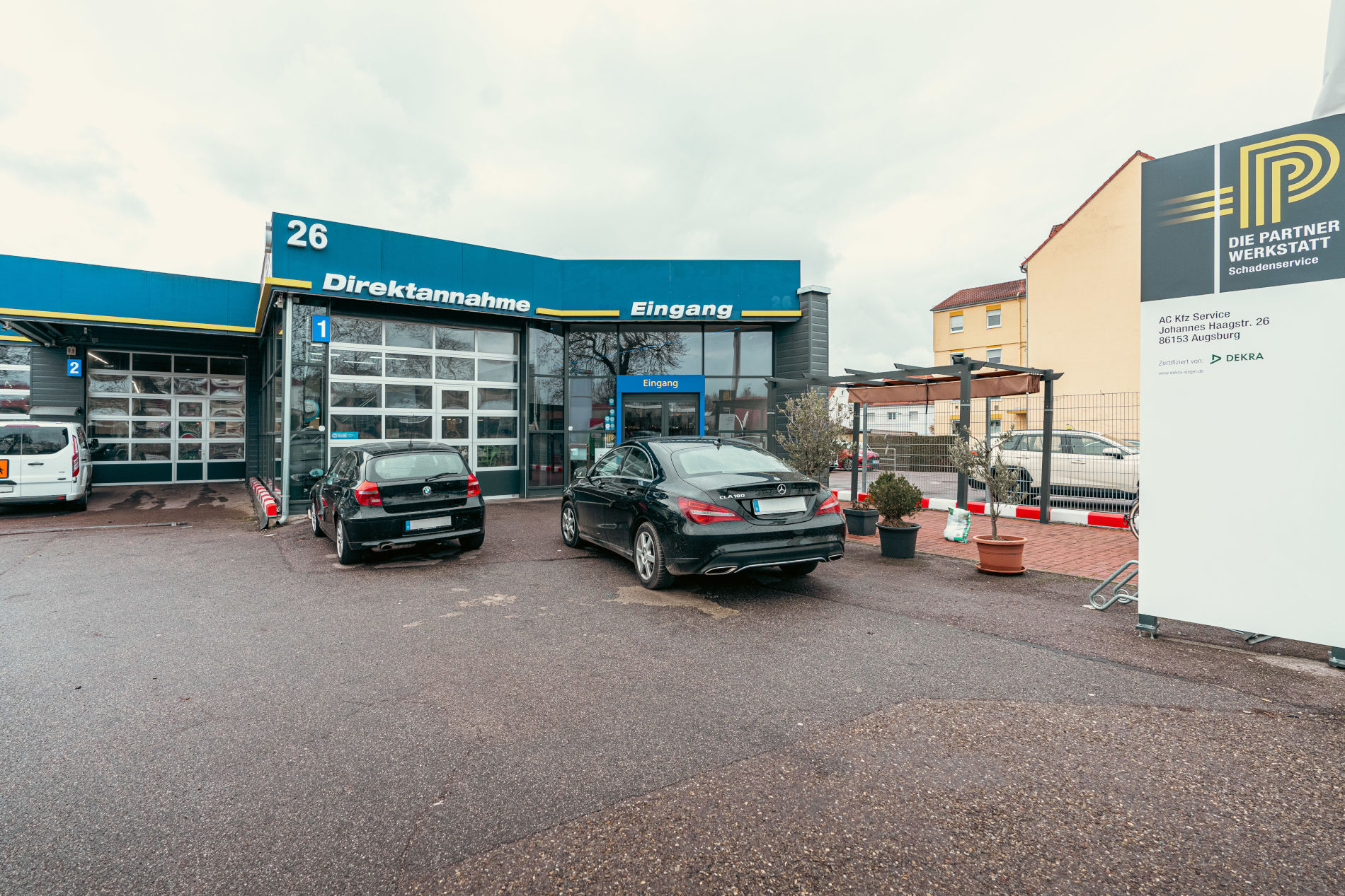 Außenansicht einer Autowerkstatt mit der Beschriftung „Direktannahme“ und „Eingang“, davor geparkte Fahrzeuge und ein Hinweisschild zur Partnerwerkstatt.